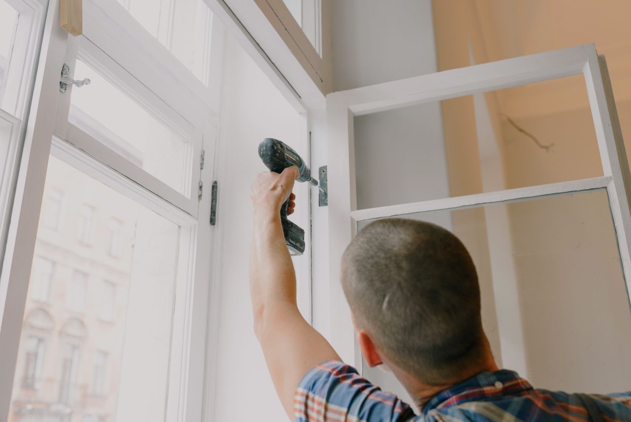 Home Back view of unrecognizable craftsman using cordless screwdriver and assembling window while screwing metal hinge during renovation works in apartment